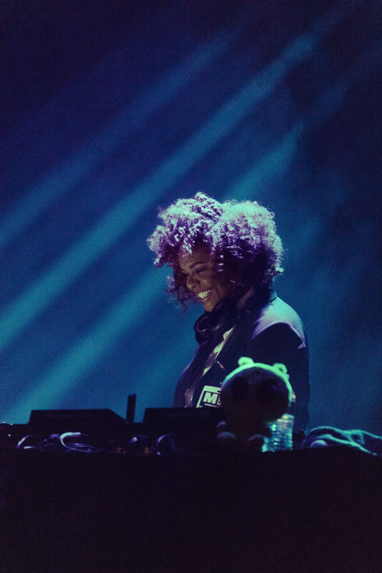 Person with curly hair smiling, standing behind DJ equipment, illuminated by blue stage lights, in a dark setting.