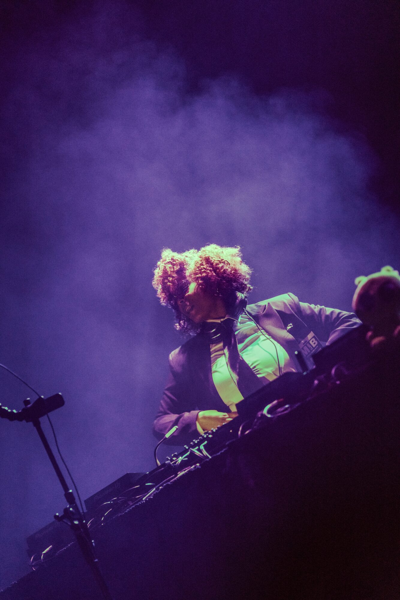 Person with curly hair DJing on stage, surrounded by purple smoke, wearing a jacket and neon green shirt, with DJ equipment.