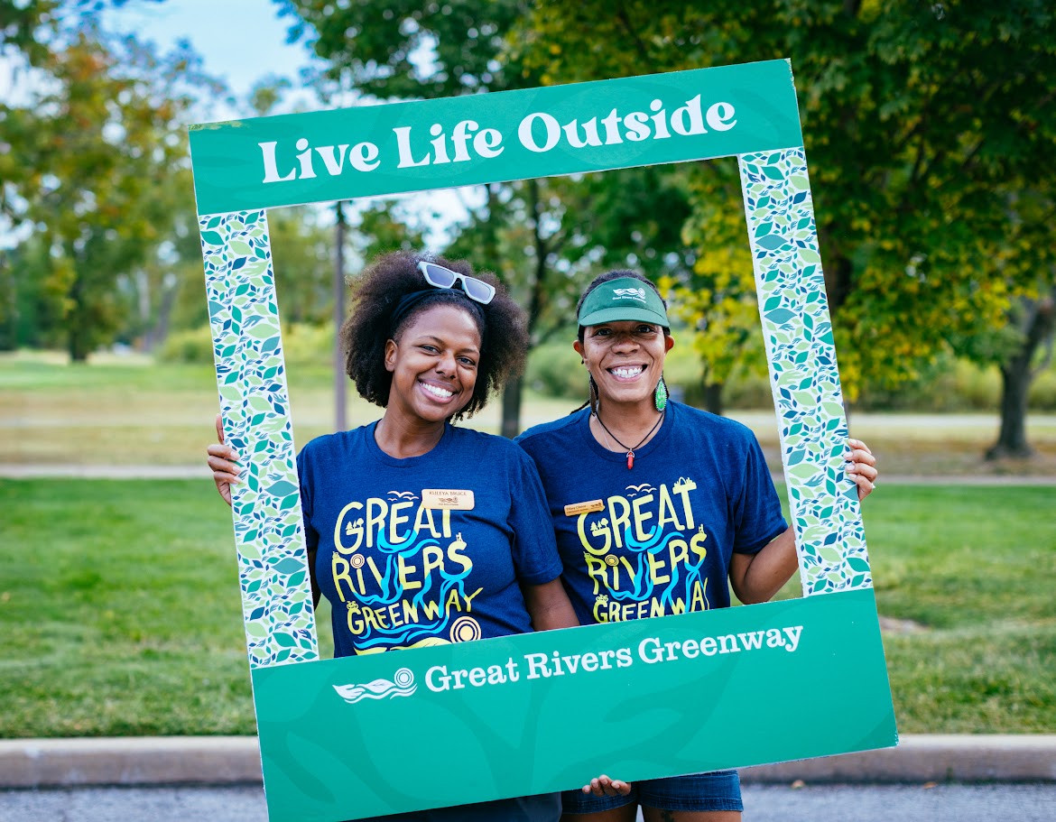 Two women smiling and holding a large photo frame outdoors, with trees and grass in the background.
