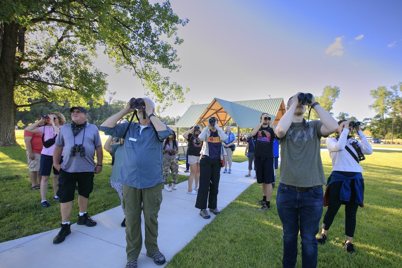 Group of people outdoors taking photos with cameras, some wearing hats, in a park with trees and tents in background.