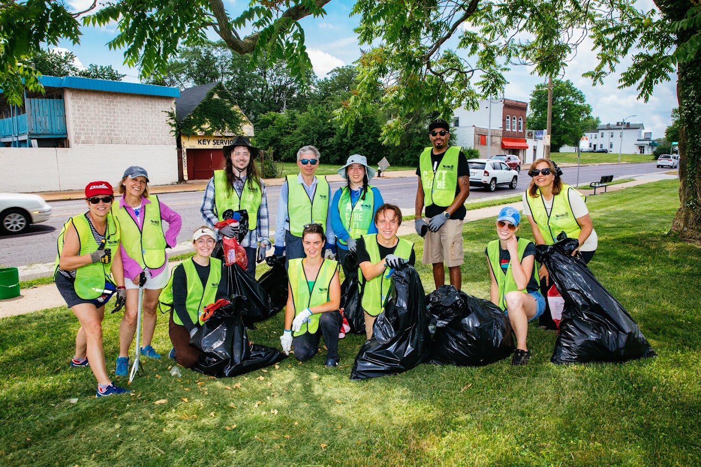 Group of ten people wearing reflective vests holding trash bags outdoors on a grassy area, with trees and buildings in the background.