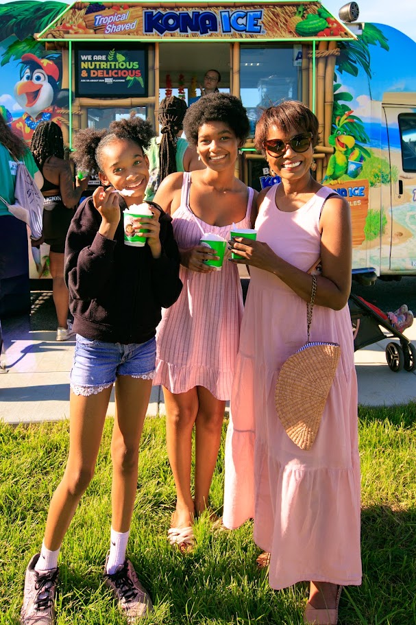 Three smiling people holding green cups in front of a Kona Ice truck at an outdoor event, sunny day.