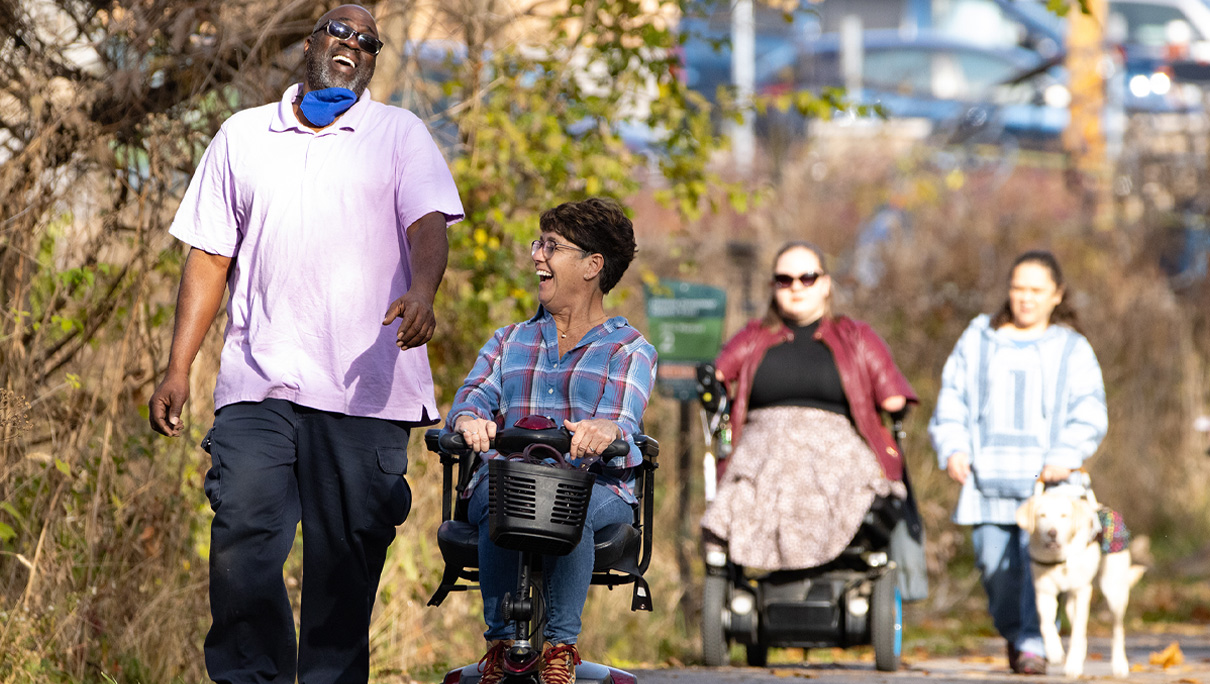 Group of people walking outdoors, including a person in a wheelchair and a person smiling at a child in a plaid shirt.