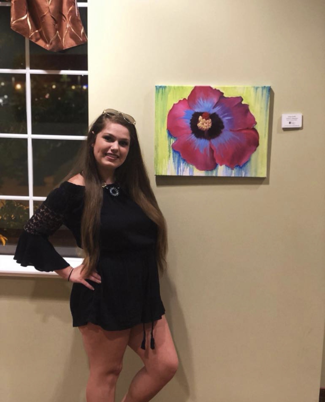 Young woman with long hair in a black dress standing next to a colorful flower painting on a wall, indoors at night.