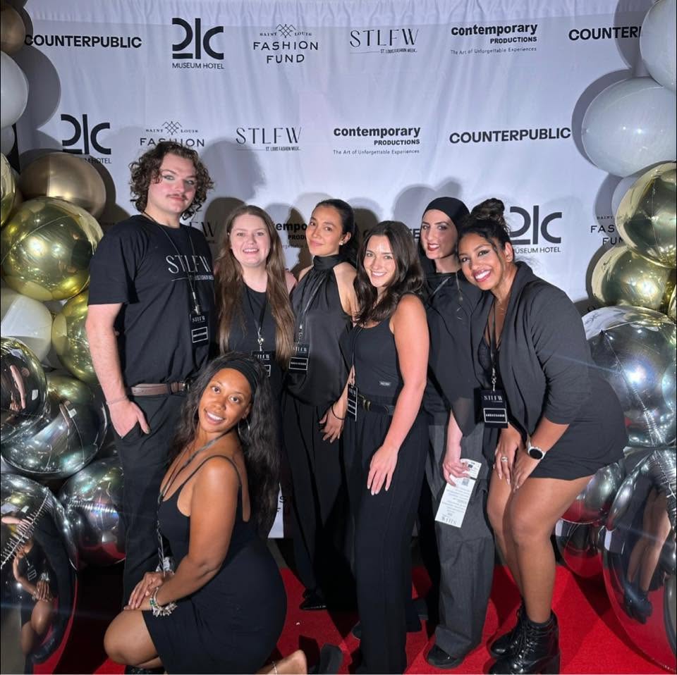 Group of seven young women posing on red carpet with balloons and backdrop, smiling and dressed stylishly.