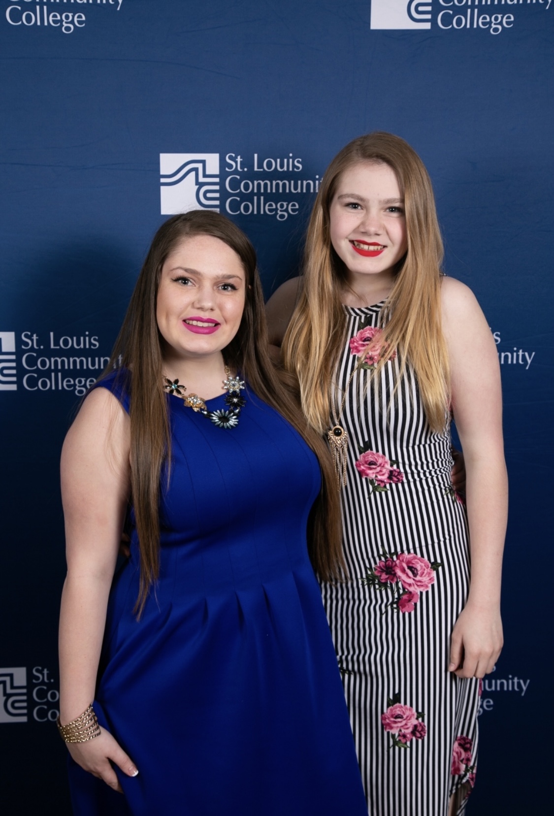 Two young women standing in front of a blue backdrop with St. Louis Community College logo, smiling, one in a blue dress, the other in a striped dress with pink flowers.