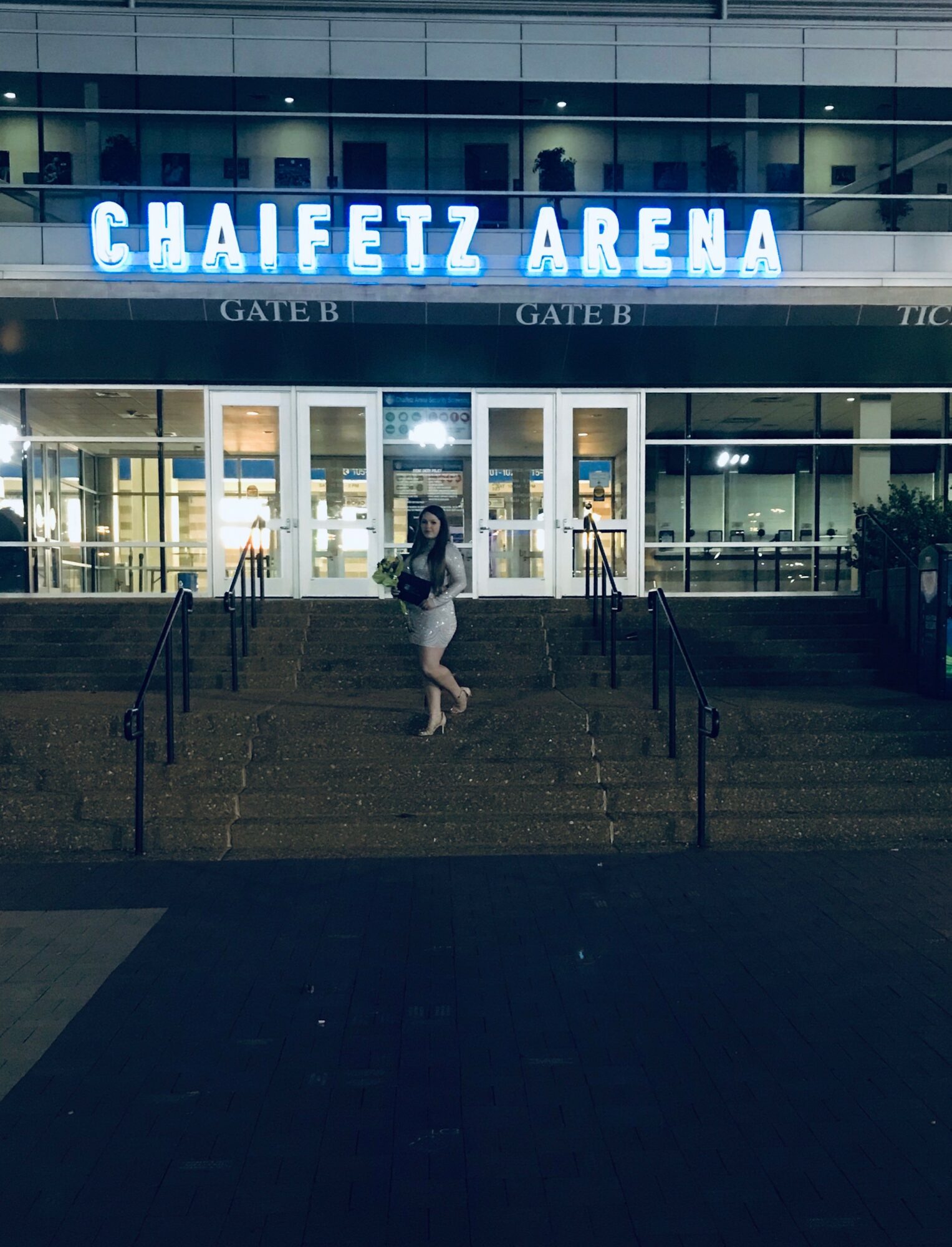 Person standing on stairs outside of Chaifetz Arena at night, illuminated sign above, glass doors behind.