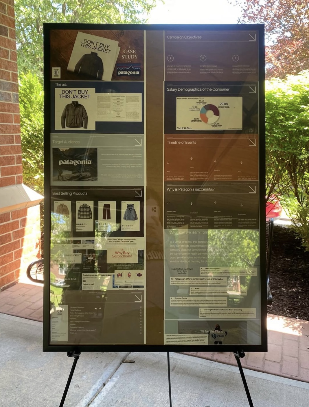 Outdoor display board with information and advertisements, divided into two sections, with trees and a brick wall in the background.