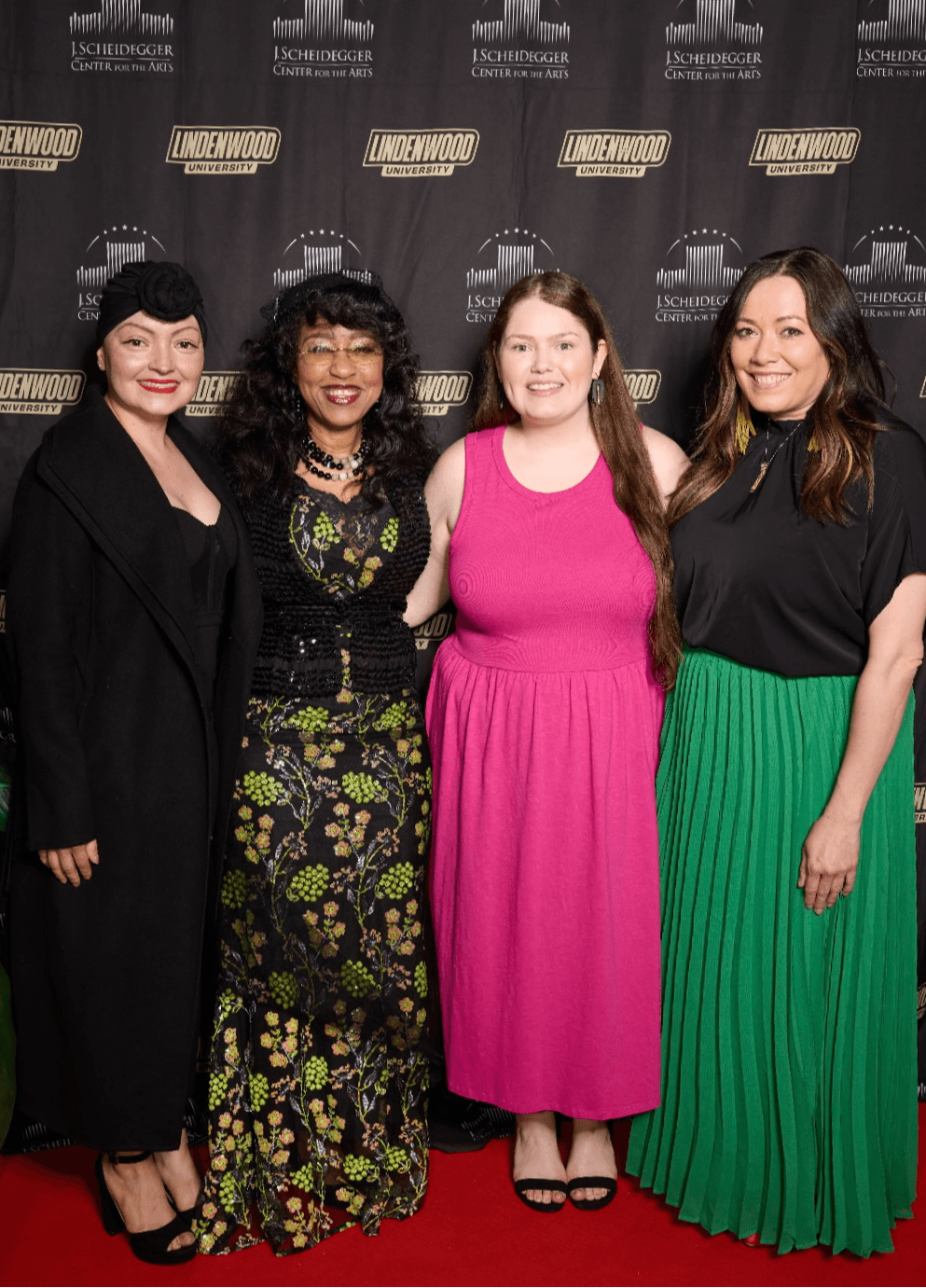 Four women standing together on a red carpet in front of a black backdrop with logos, smiling for the photo.