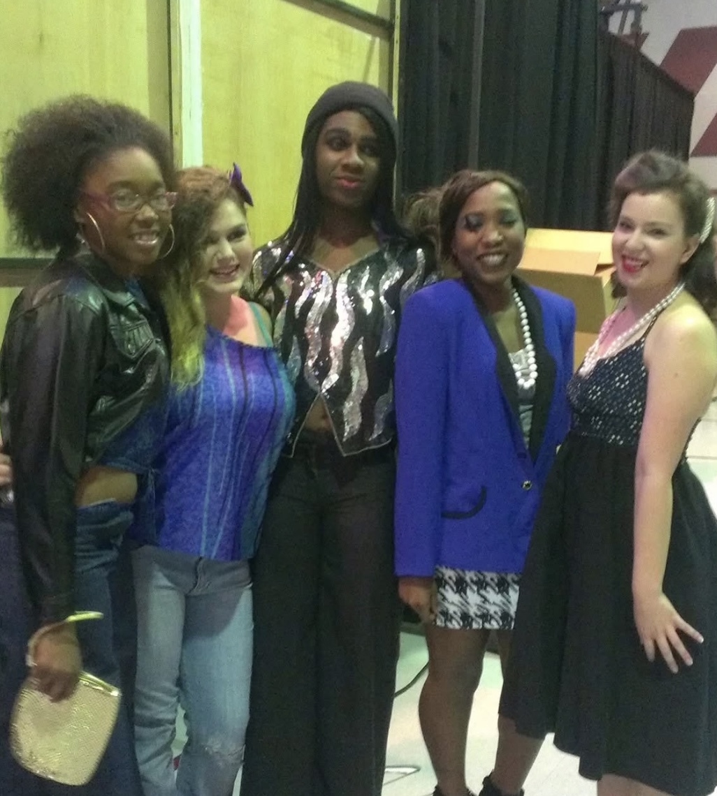 Five women standing together, smiling, indoors with green and dark curtains in the background.