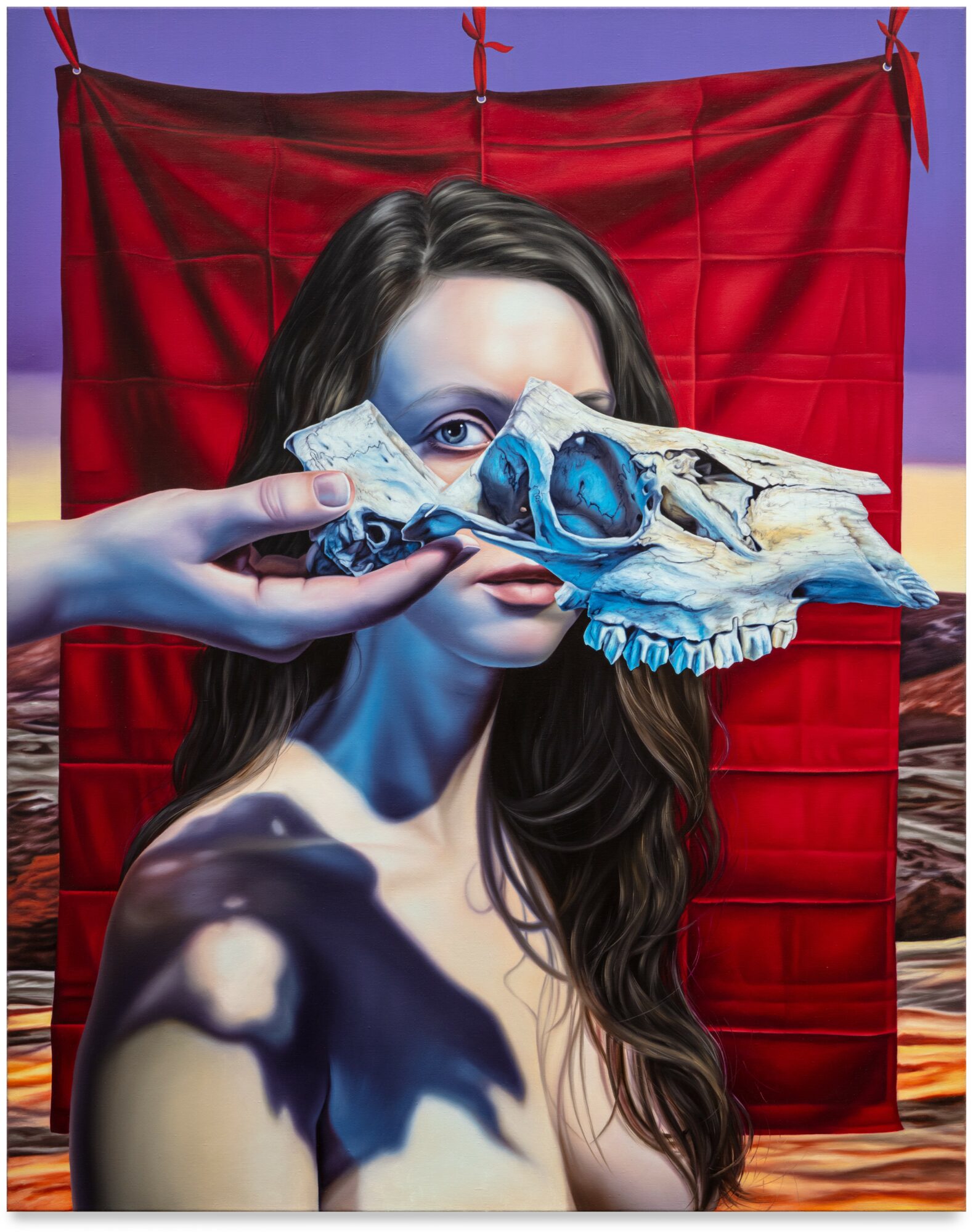 A woman with long hair holds a skull with blue eyes in front of her face, with a red cloth background.