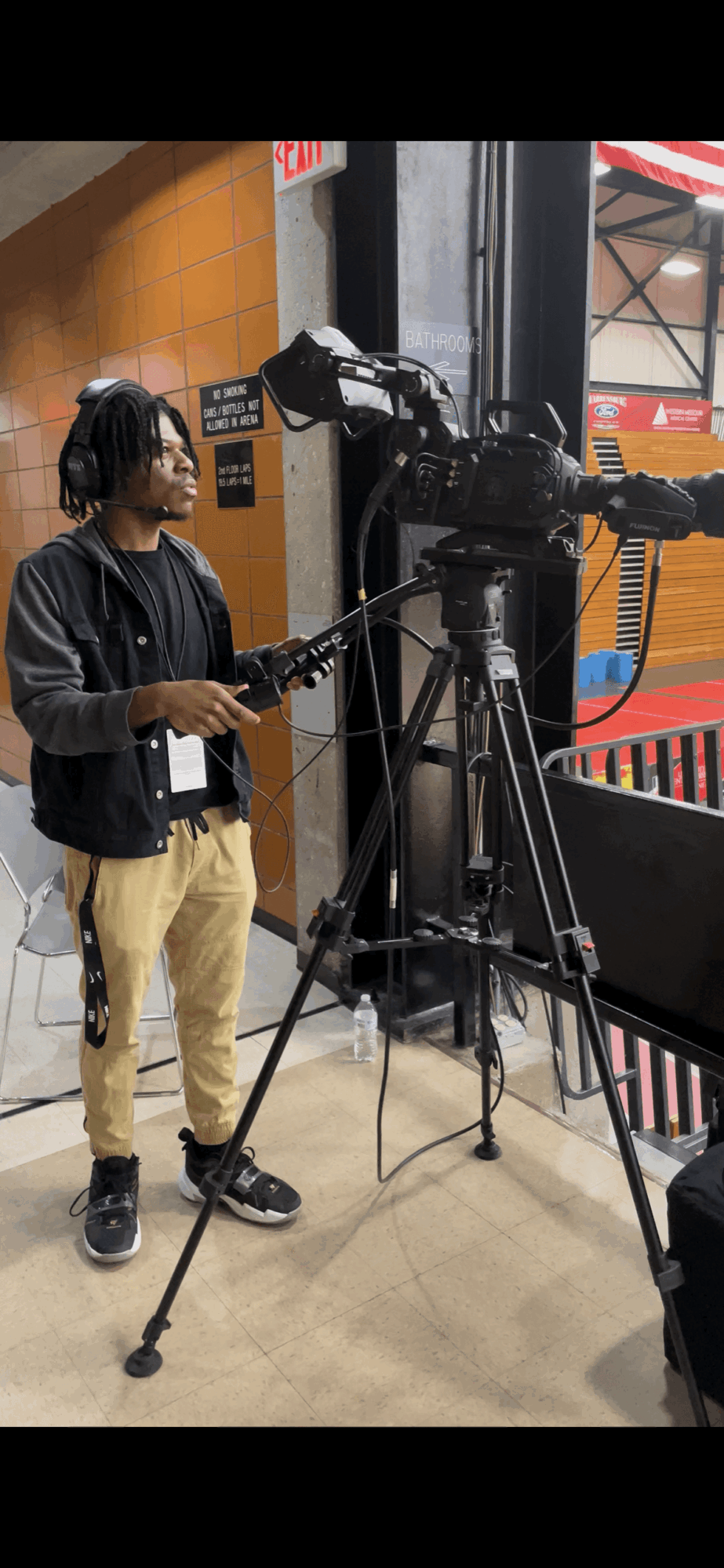 Person operating a professional video camera on a tripod indoors, wearing headphones and casual clothing.