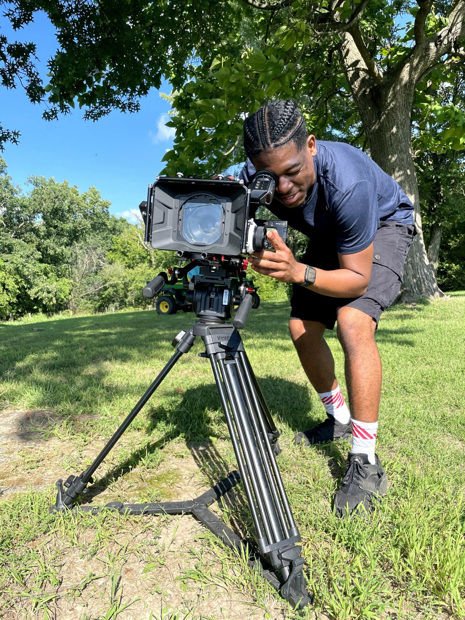 Person operating a professional video camera outdoors on grass with trees and blue sky in background.