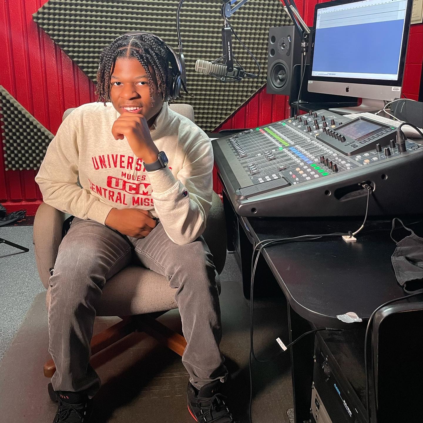 Child with braided hair sitting in a recording studio, smiling, with audio equipment and a computer screen behind him.