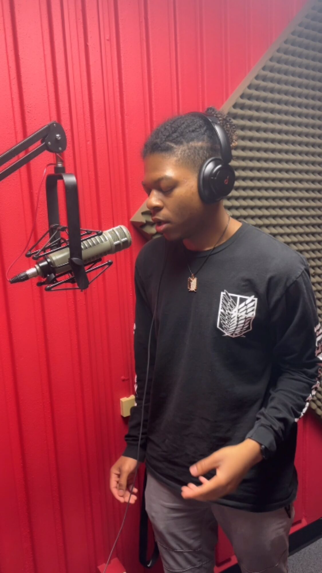 Young person with headphones speaking into a microphone in a recording studio, red wall background.