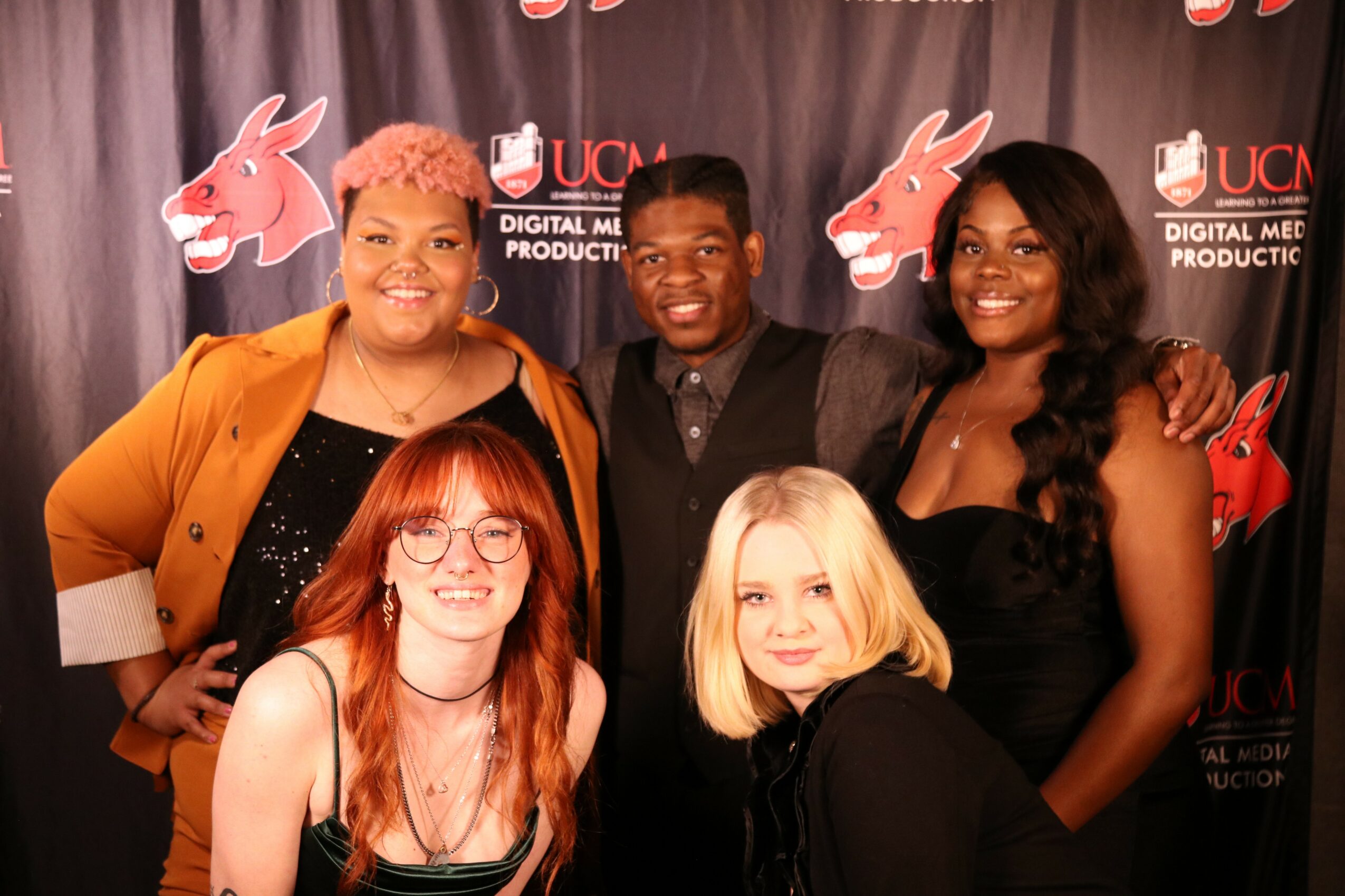 Group of five people smiling in front of a black backdrop with red and white logos, some with arms around each other.