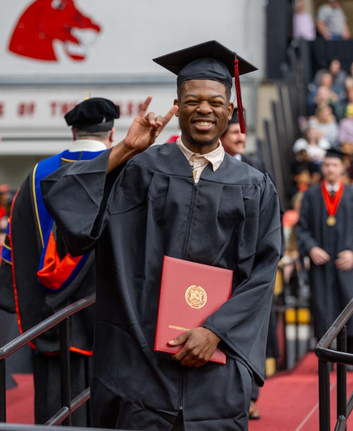 Graduate in cap and gown smiling and making a peace sign, holding a diploma, at graduation ceremony.