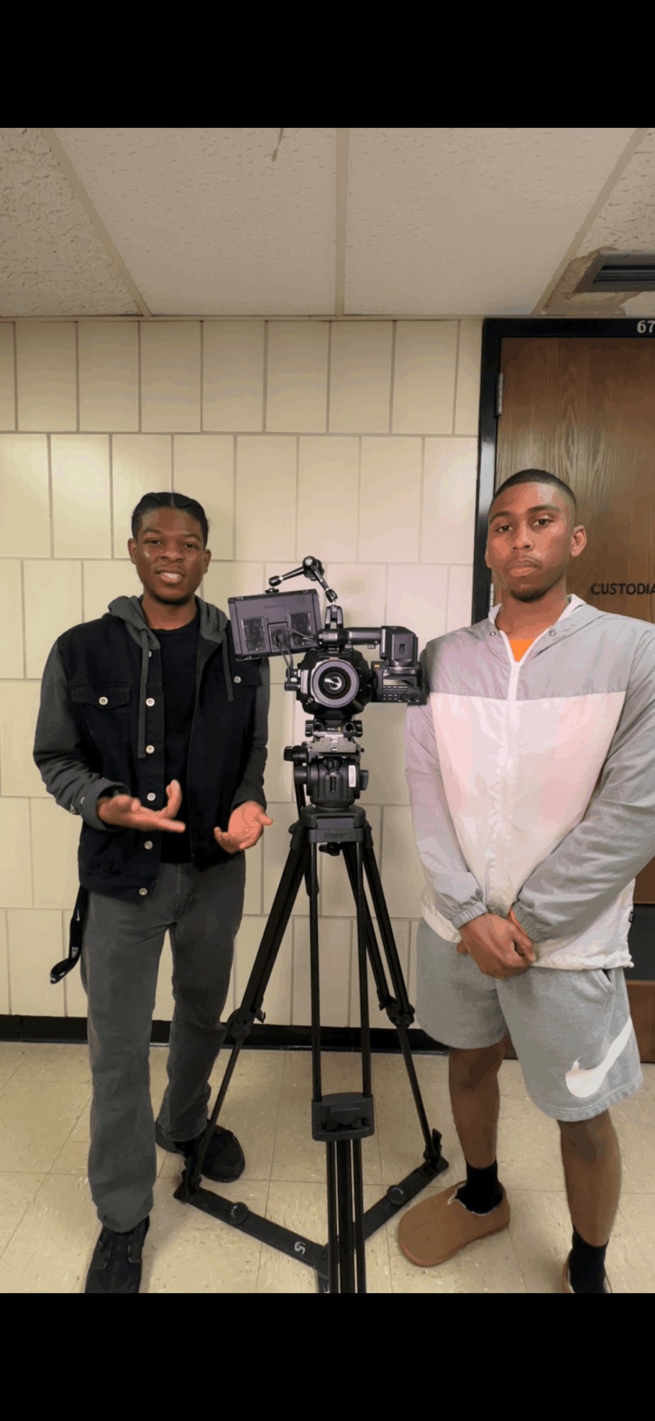 Two young men stand behind a camera on a tripod in a room with beige tiled walls and a wooden door.