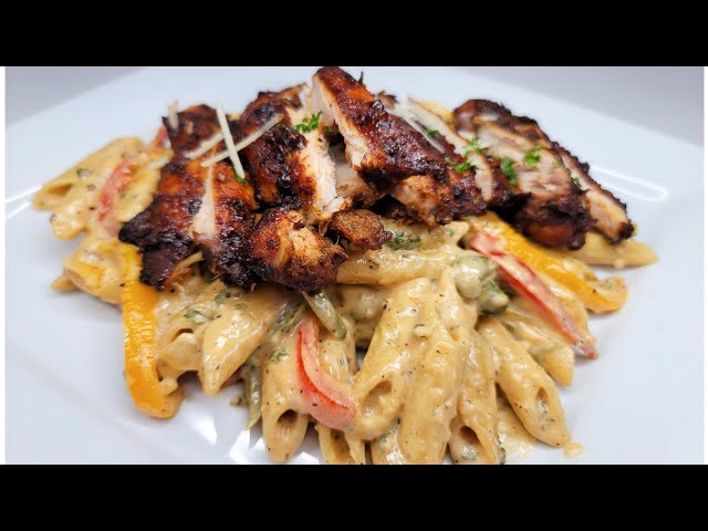 Grilled chicken pieces served over creamy pasta with vegetables on a white plate.
