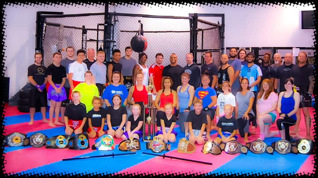 Group of children and adults in a gym with boxing equipment, some holding trophies, colorful mats on the floor.