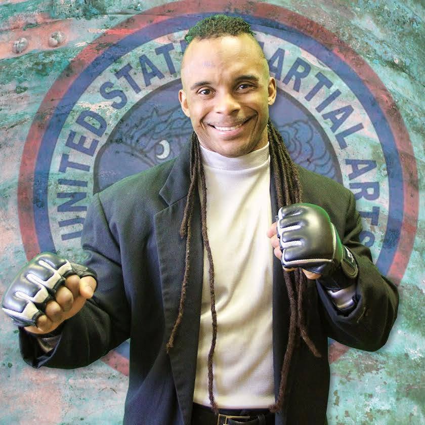 Person with long dreadlocks wearing a black jacket and gloves, smiling with fists raised, in front of a circular logo background.