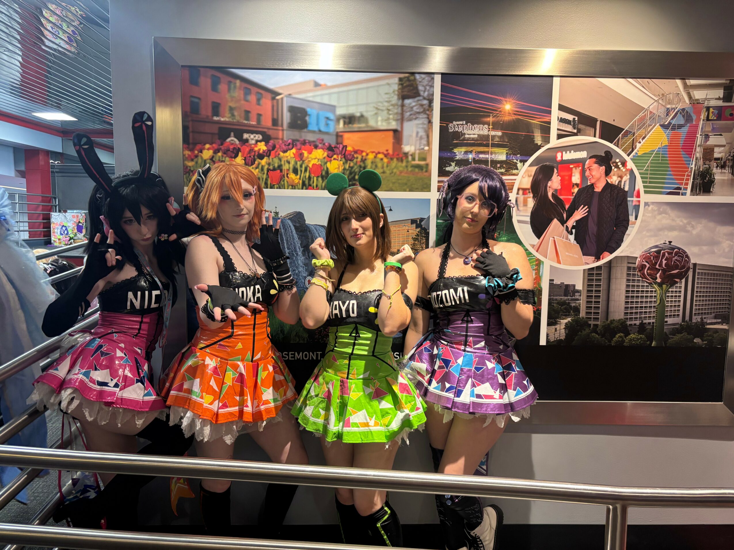 Four women in colorful costumes pose in front of a cityscape display, with flowers and city buildings in the background.