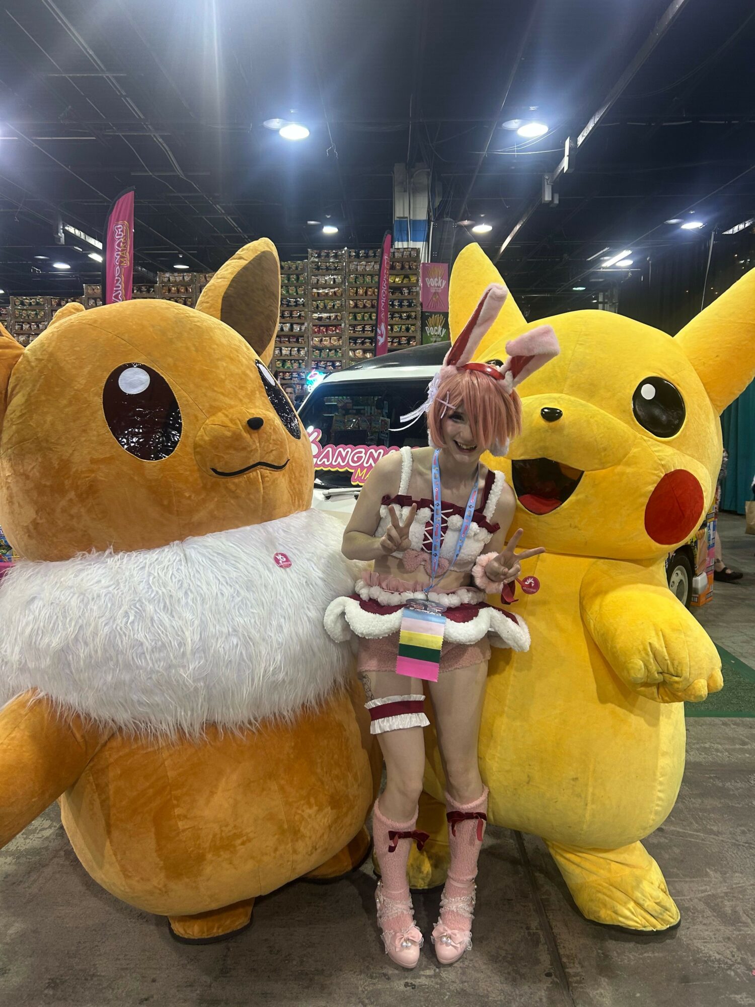 Person in a Pikachu costume with a large yellow body and red cheeks, standing next to a large Eevee plush, in a store or event space.