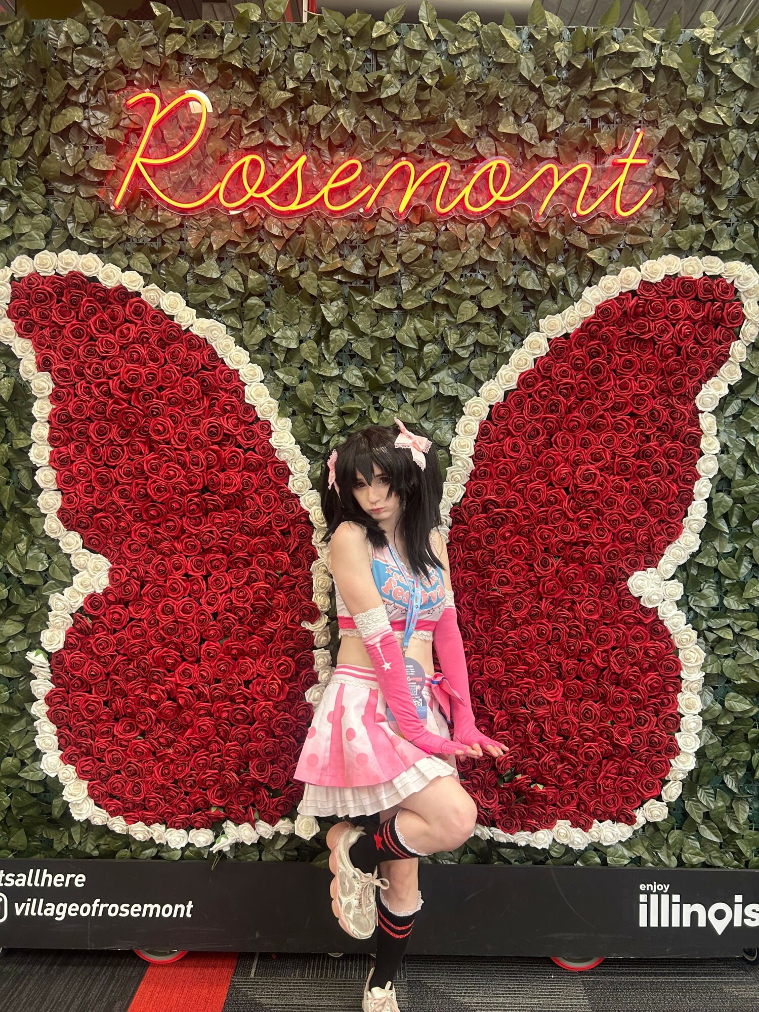 Girl in pink dress sitting on a bench with large red and white heart-shaped flower arrangements behind her, and a neon sign reading 'Reverence'.