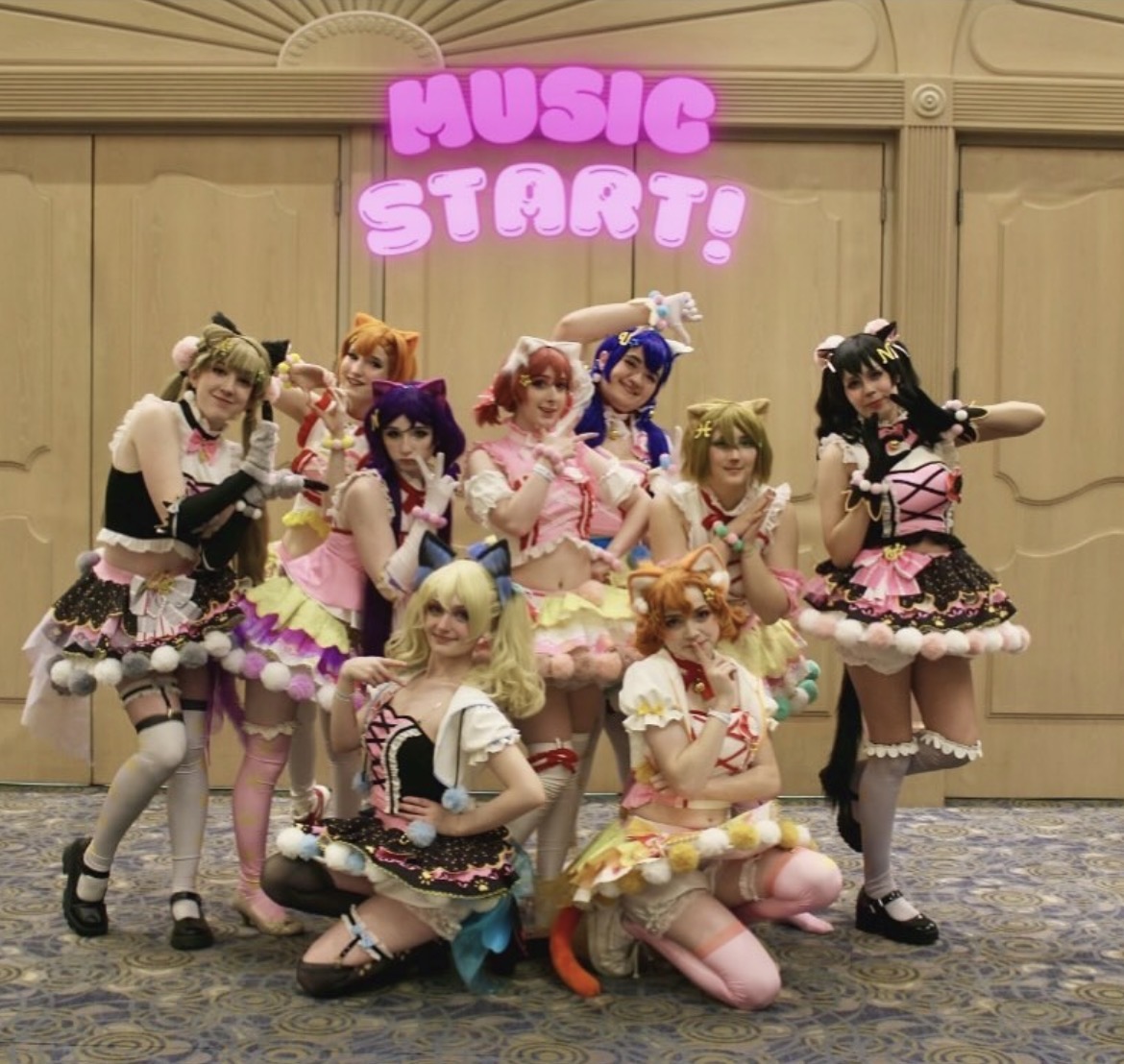 Group of ten people in colorful costumes posing indoors with a pink sign that says 'Music Start!'.