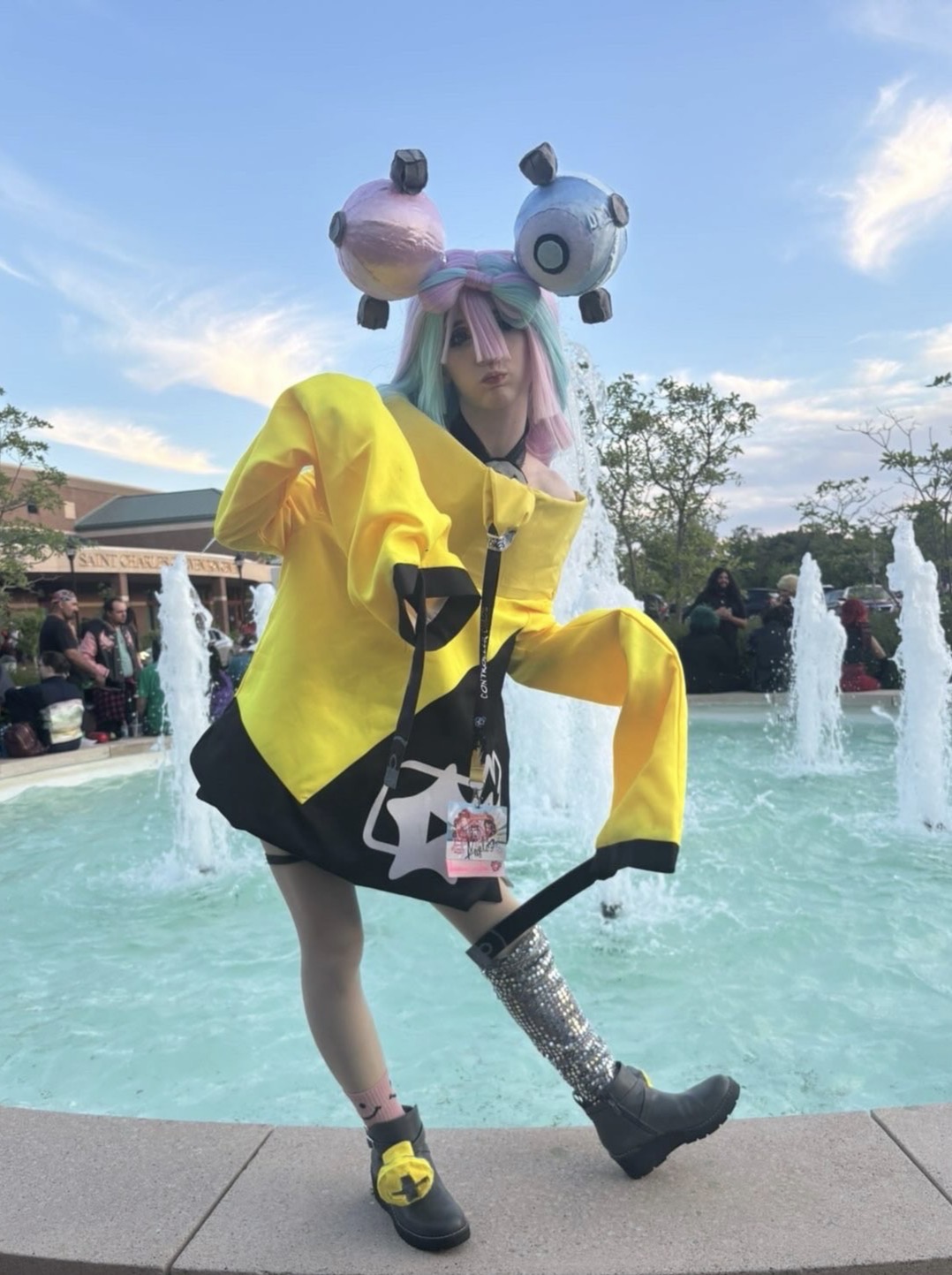 Person standing on a fountain edge wearing a yellow jacket, holding an umbrella, with a balloon hat, in an outdoor park.
