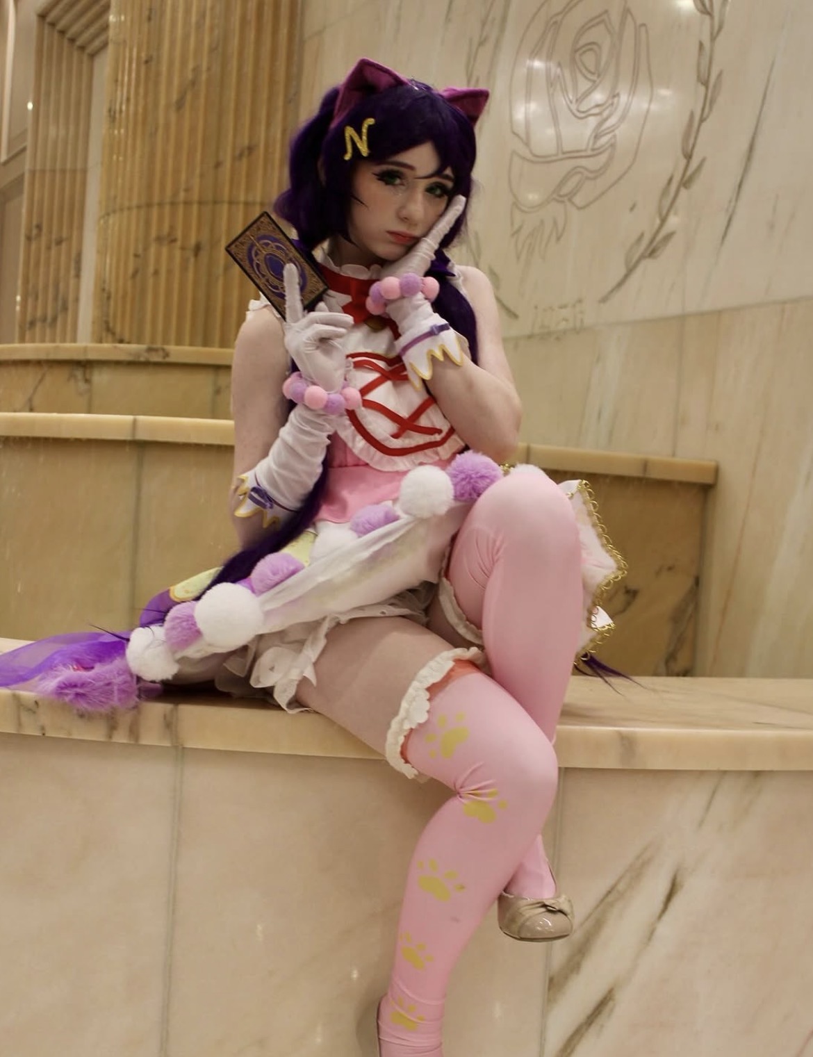 Person in colorful costume with purple hair, sitting on a wooden surface, holding a card and a plush toy, with a wooden wall background.