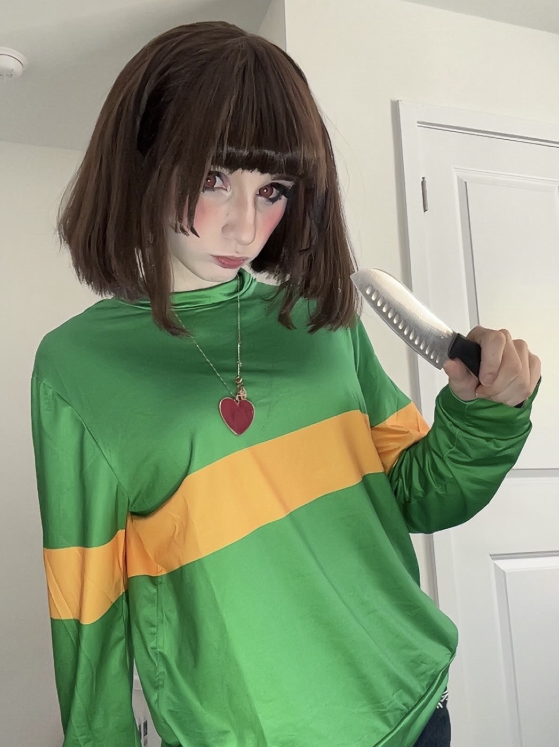 Young woman with shoulder-length brown hair and bangs, wearing a green and yellow striped top, holding a hairbrush, standing indoors.