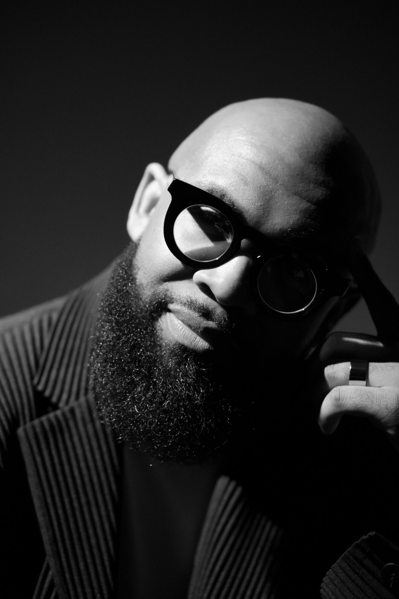 Bald person with glasses, head tilted, hand near face, in black and white, wearing a striped suit.