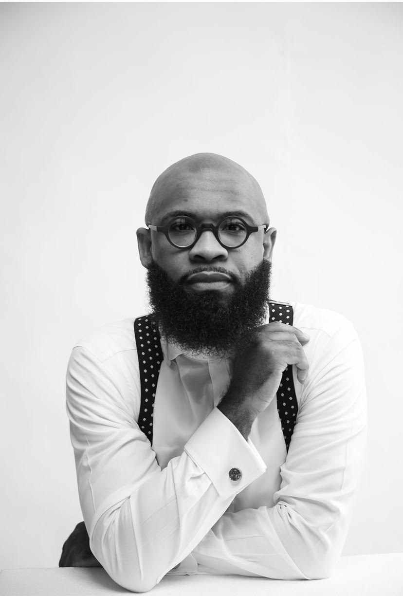 Man with glasses and a beard, wearing a white shirt and suspenders, resting his chin on his hand.