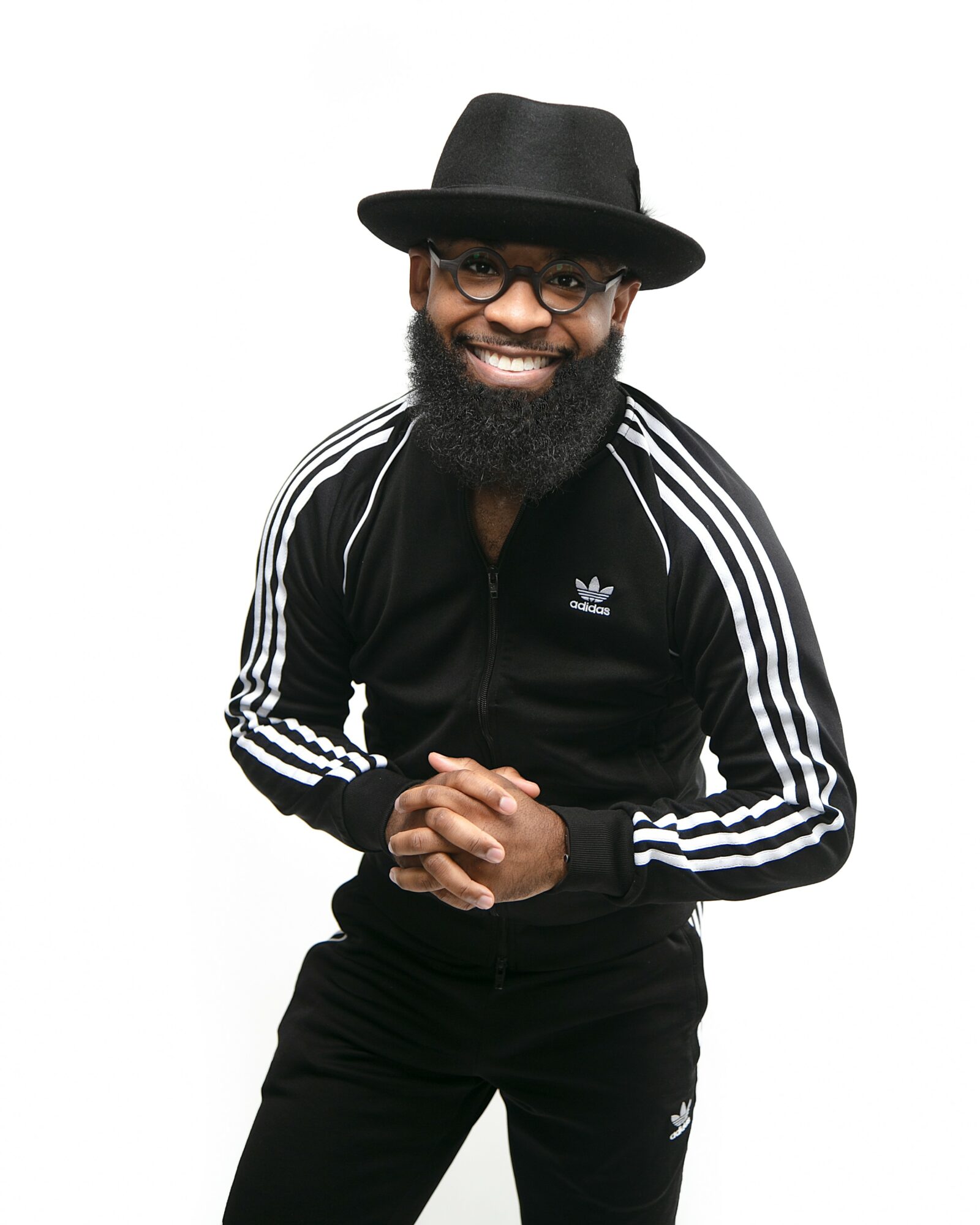 Smiling man with glasses, beard, black hat, black Adidas jacket, and hands clasped, standing against a white background.