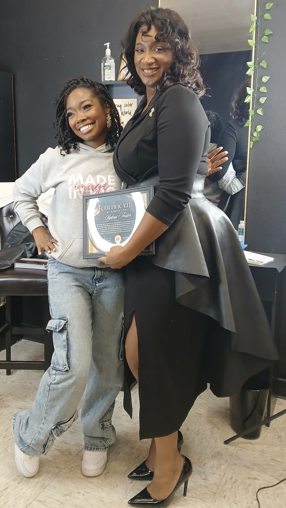 Two women smiling, one holding a framed picture, standing indoors with a dark wall and green plant in background.