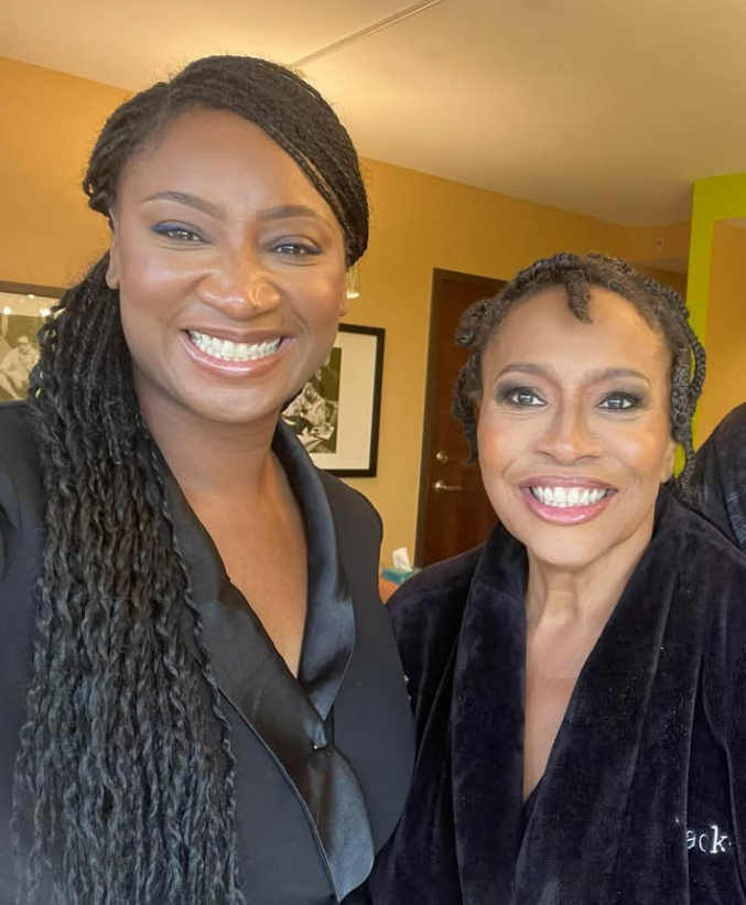 Two women smiling indoors, one with long braided hair and the other with short curly hair, both wearing dark clothing.