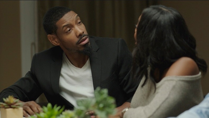 Two people sitting at a table engaged in conversation, with plants in the foreground.