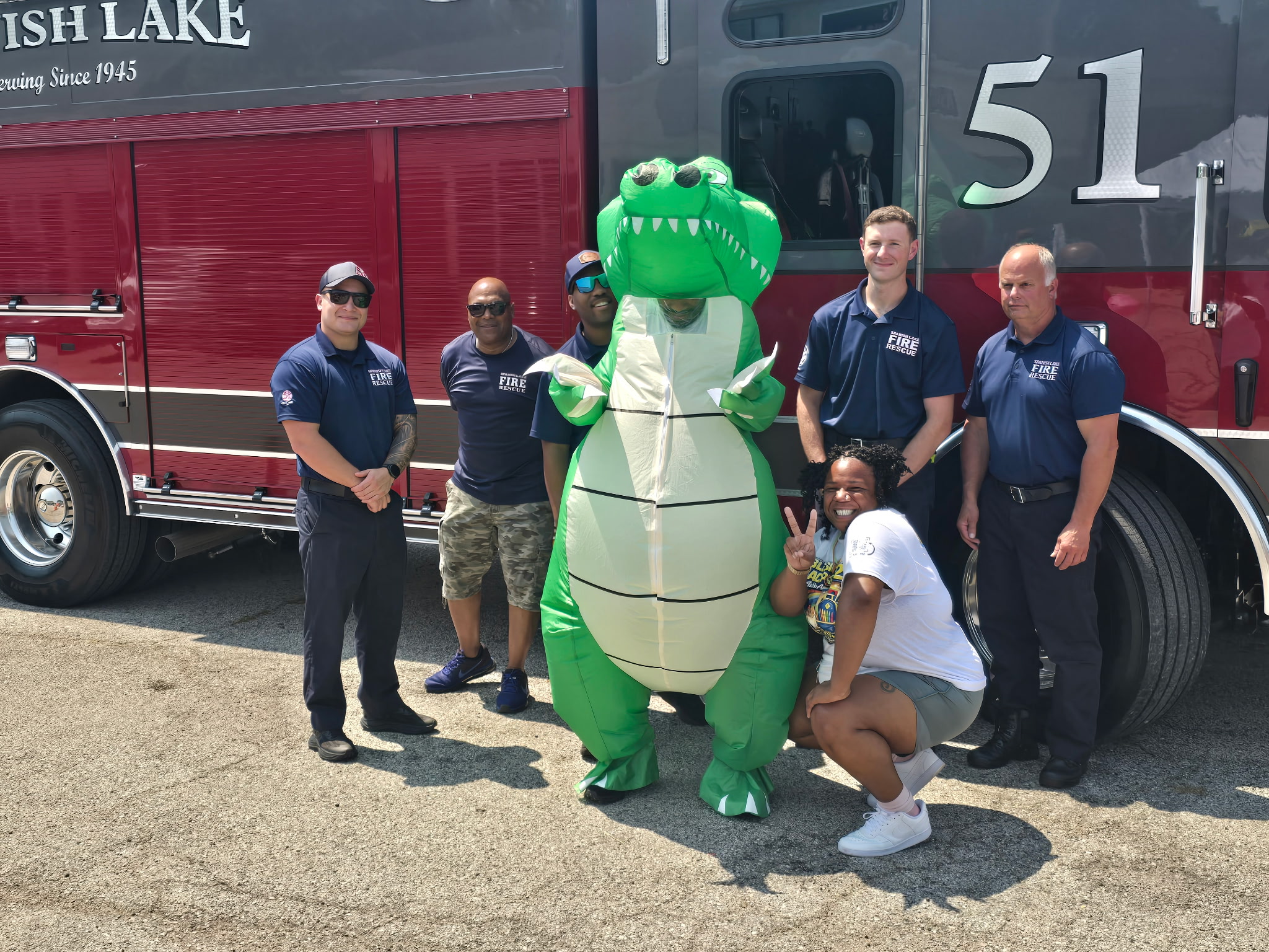 Group of five people and a person in a green alligator costume standing in front of a fire truck, smiling outdoors.