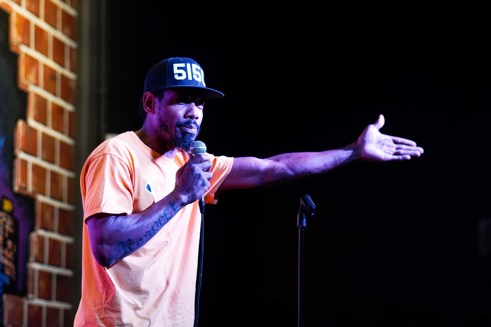 Man performing stand-up comedy on stage, holding microphone, gesturing with his left hand, wearing a cap and orange shirt.