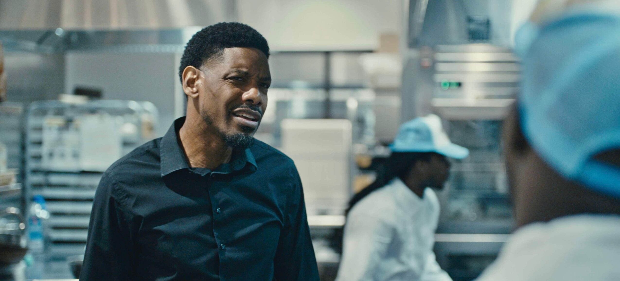 Man smiling and talking to a person in a kitchen or food preparation area, with workers in the background.