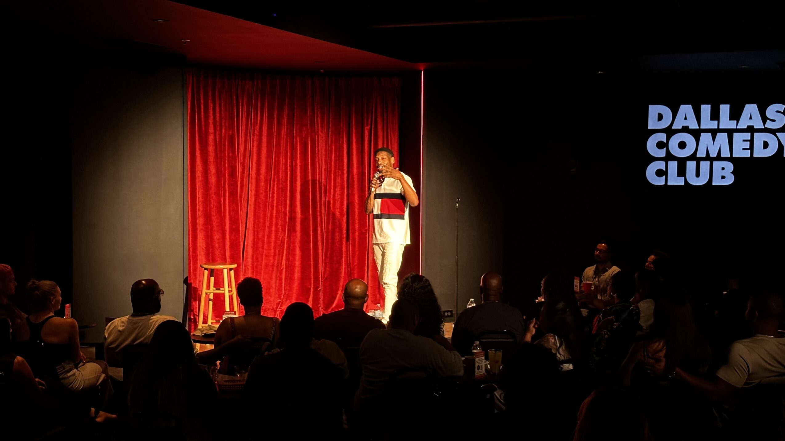 Stand-up comedian on stage with red curtain, audience seated, Dallas Comedy Club sign visible, dim lighting.