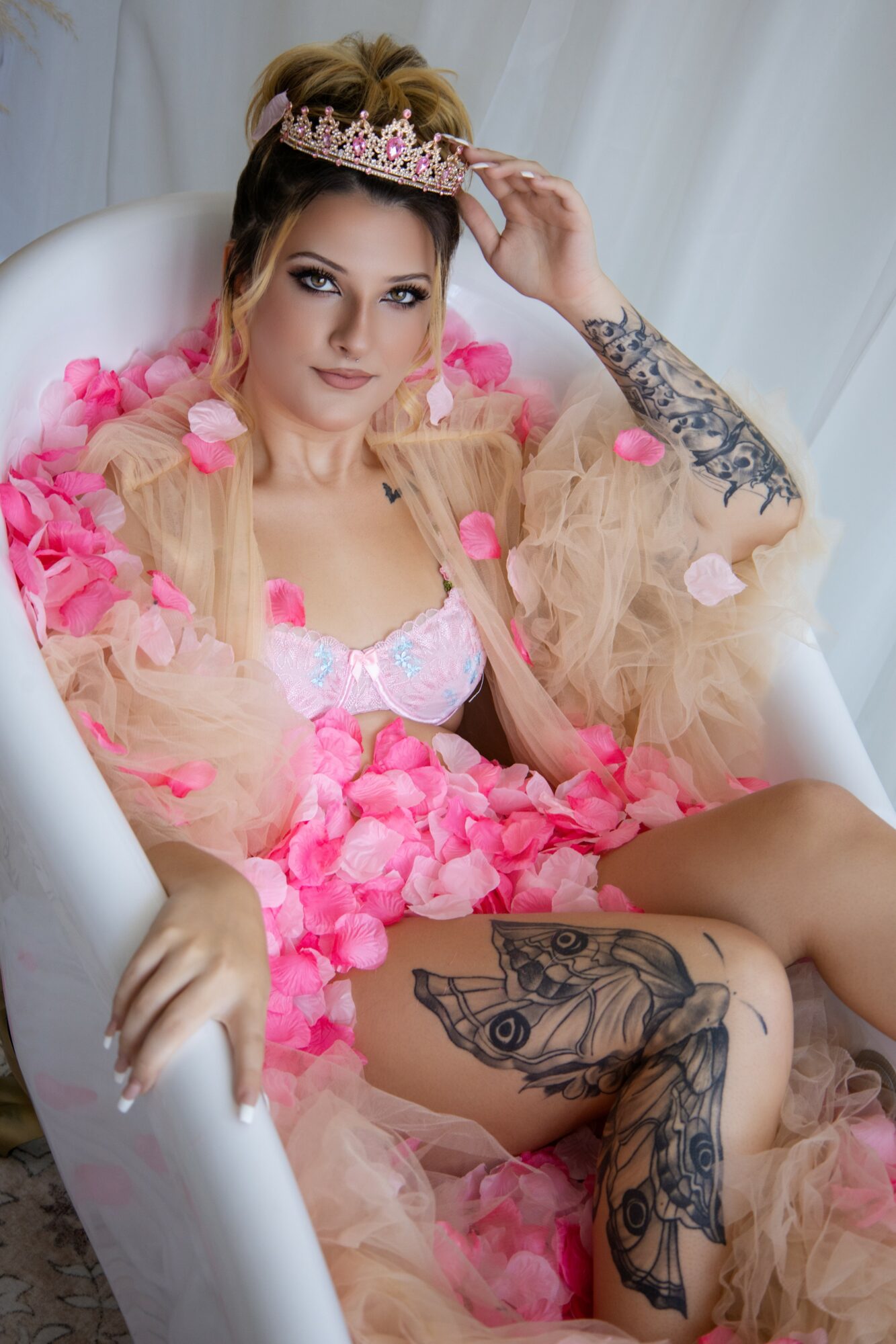 Woman with a tiara, sitting in a white chair, wearing a pink dress with pink petals, tattoos visible on arm and leg.