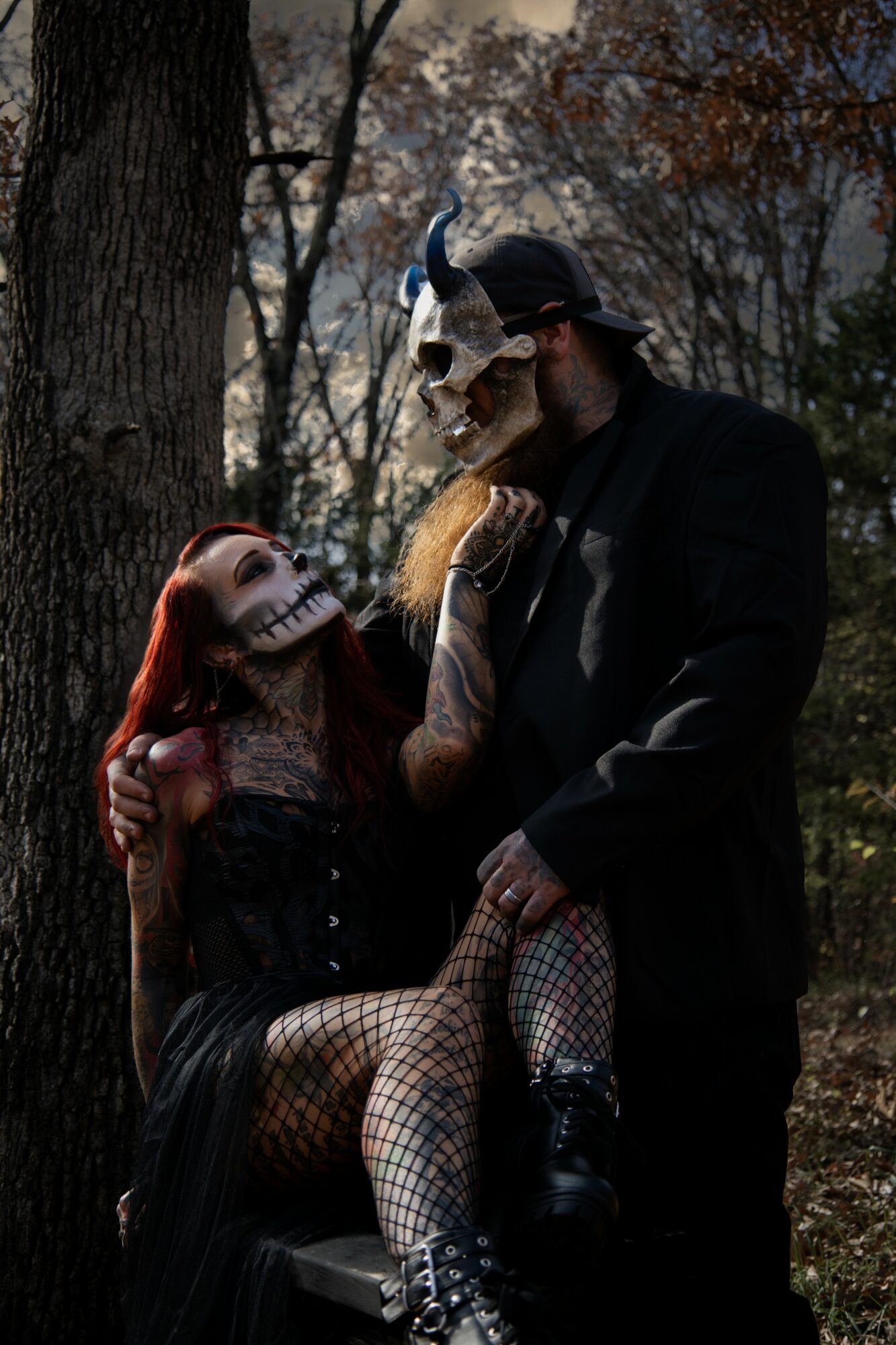 A man and a woman in Halloween costumes outdoors, with trees in the background. The woman is sitting on a box, and the man is standing, both wearing masks.