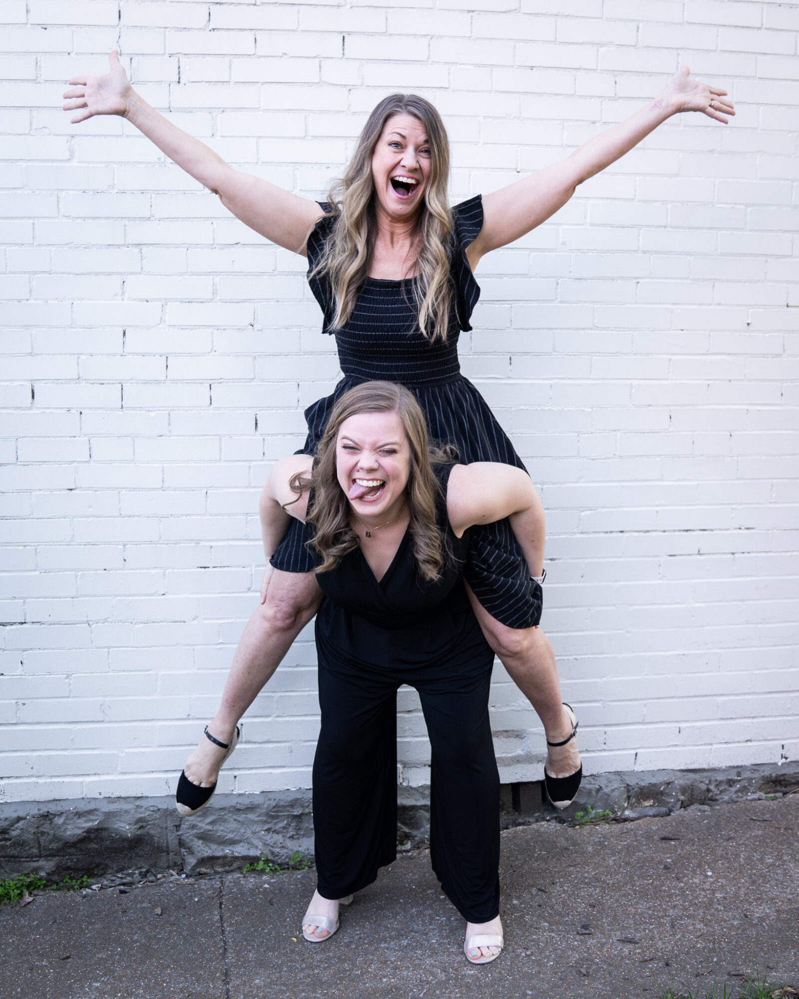 Two women smiling and laughing, one giving a piggyback ride against a white brick wall.