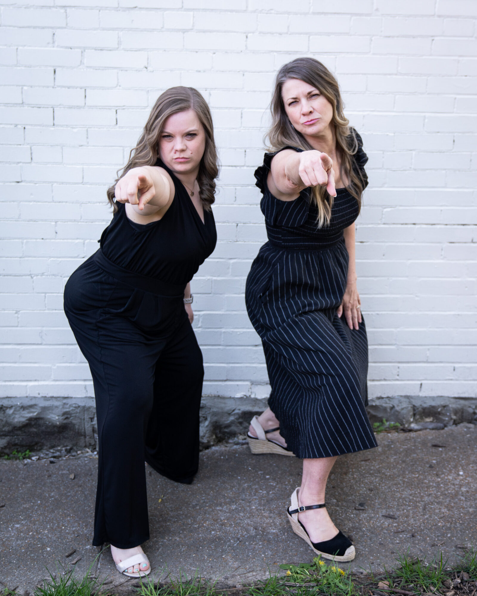 Two women stand against a white brick wall, pointing directly at the camera, with serious expressions.