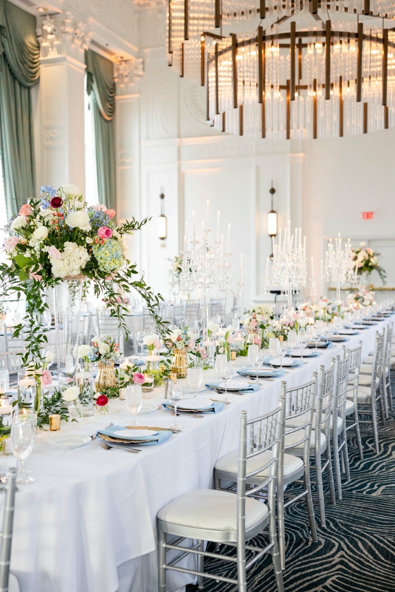 Elegant banquet table with floral centerpieces, candles, and place settings in a bright, decorated room with large windows and chandelier.