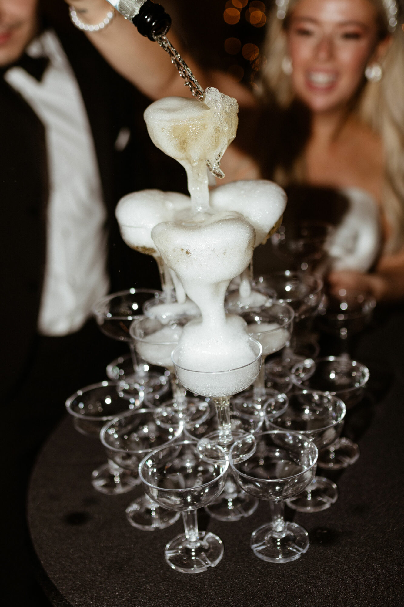 People in formal attire pour foam into a tower of glasses at a celebration, smiling and enjoying the moment.