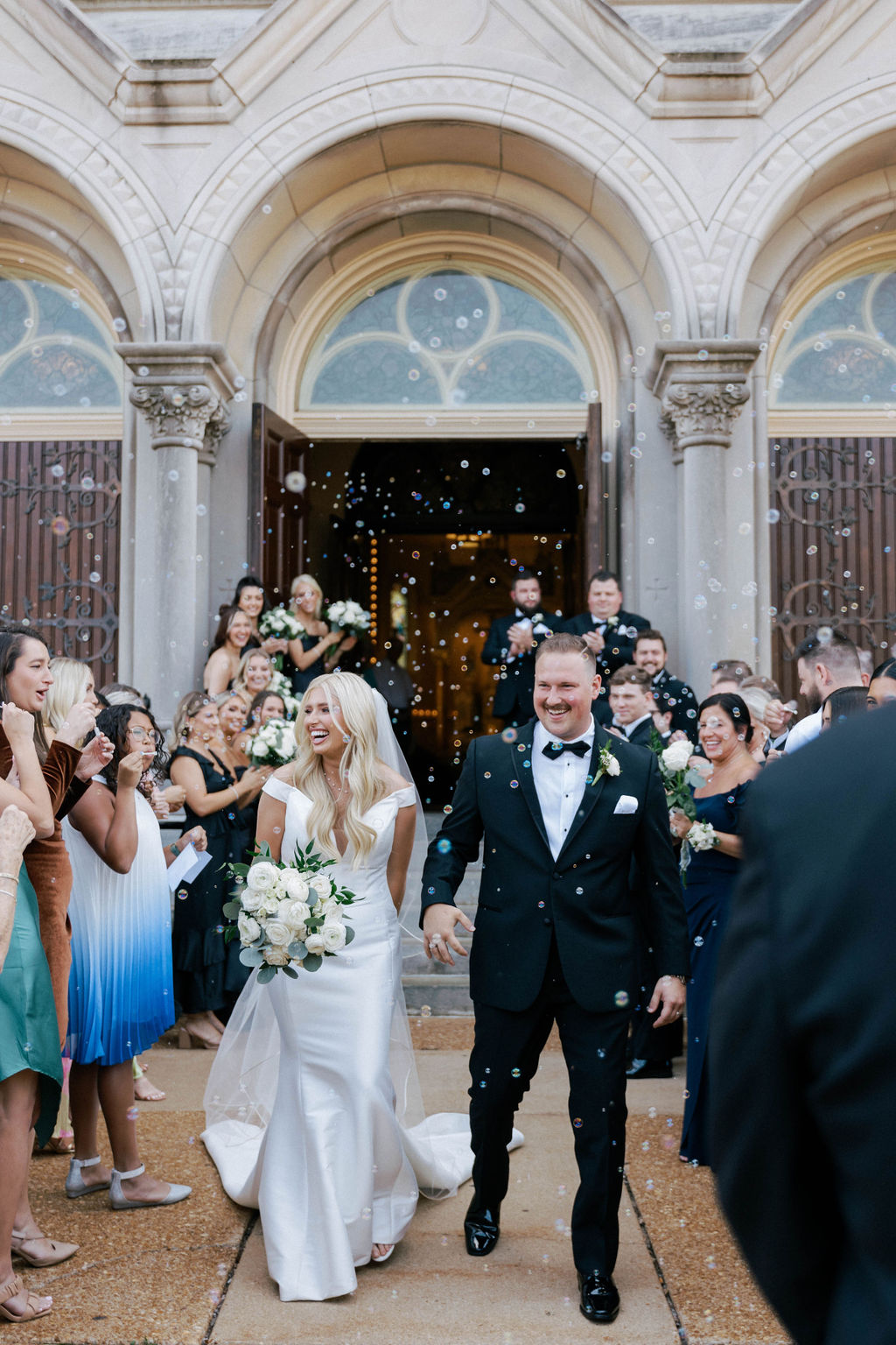 Bride and groom walk out of a building with guests celebrating and throwing confetti, ornate architecture in background.