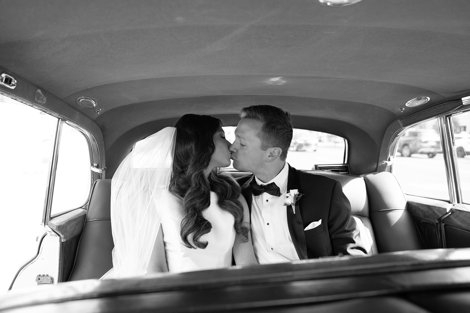 Bride and groom sitting inside a vehicle, facing each other, about to kiss, in formal wedding attire.